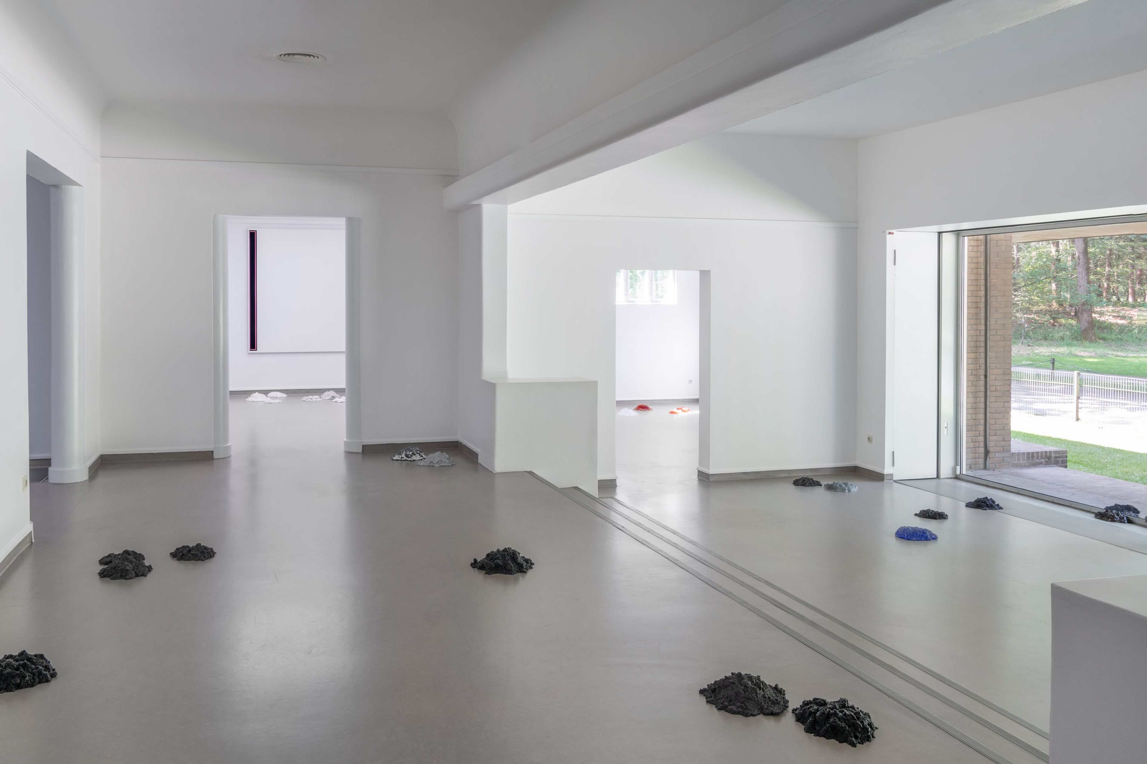 Zaalfoto Sarah van Sonsbeeck, _Moles of Modernism_ Foto van een museumzaal met verspreid over de lichtgrijze vloer verschillende (mols)hopen in allerlei kleuren en materialen. Rechts een raam met blik naar een weg, links twee deuren.