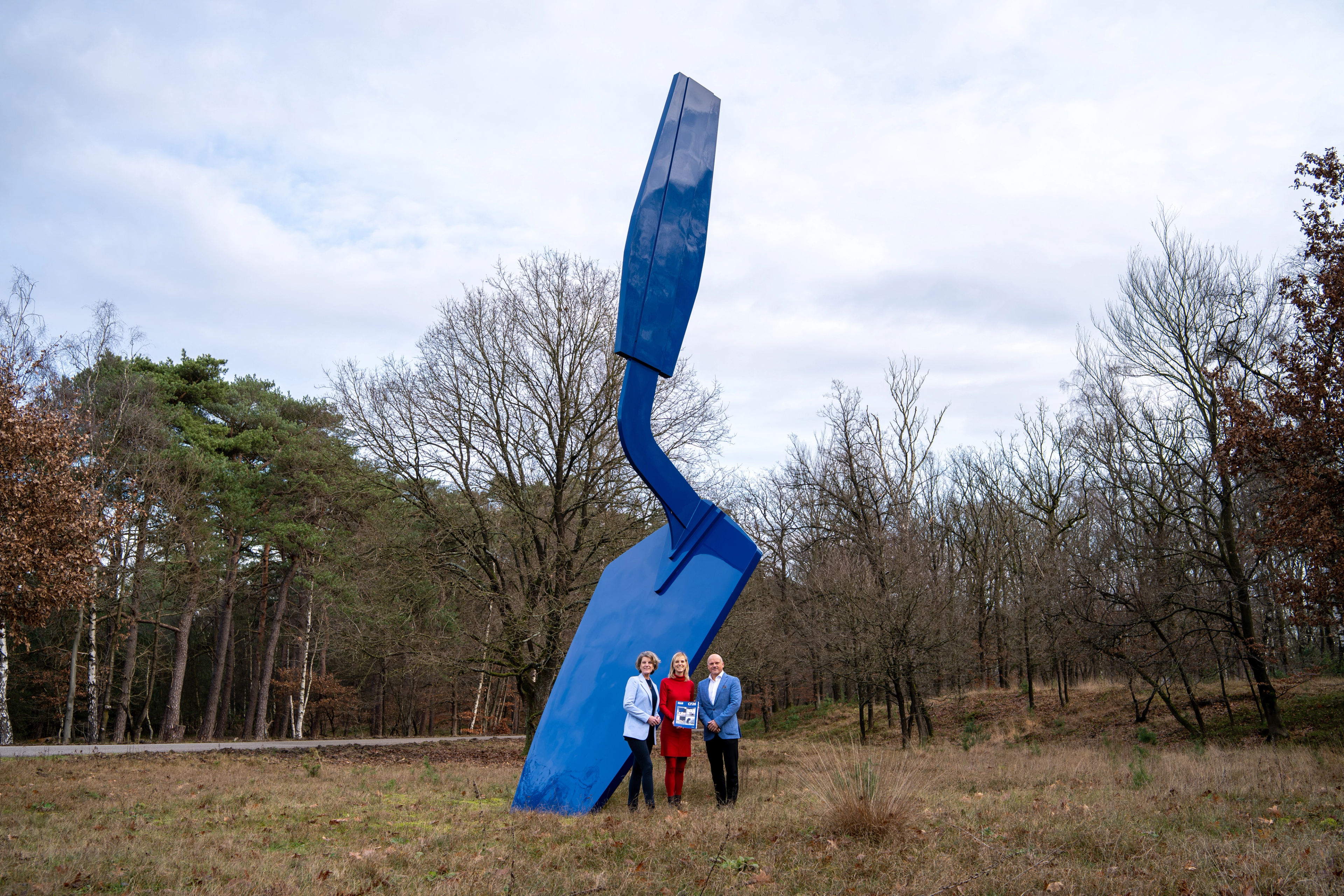 Drie personen staan bij een groot blauw kunstwerk in de vorm van een troffel, ze houden samen een soort akte vast en kijken blij de camera in. Op een open veld tussen bomen.