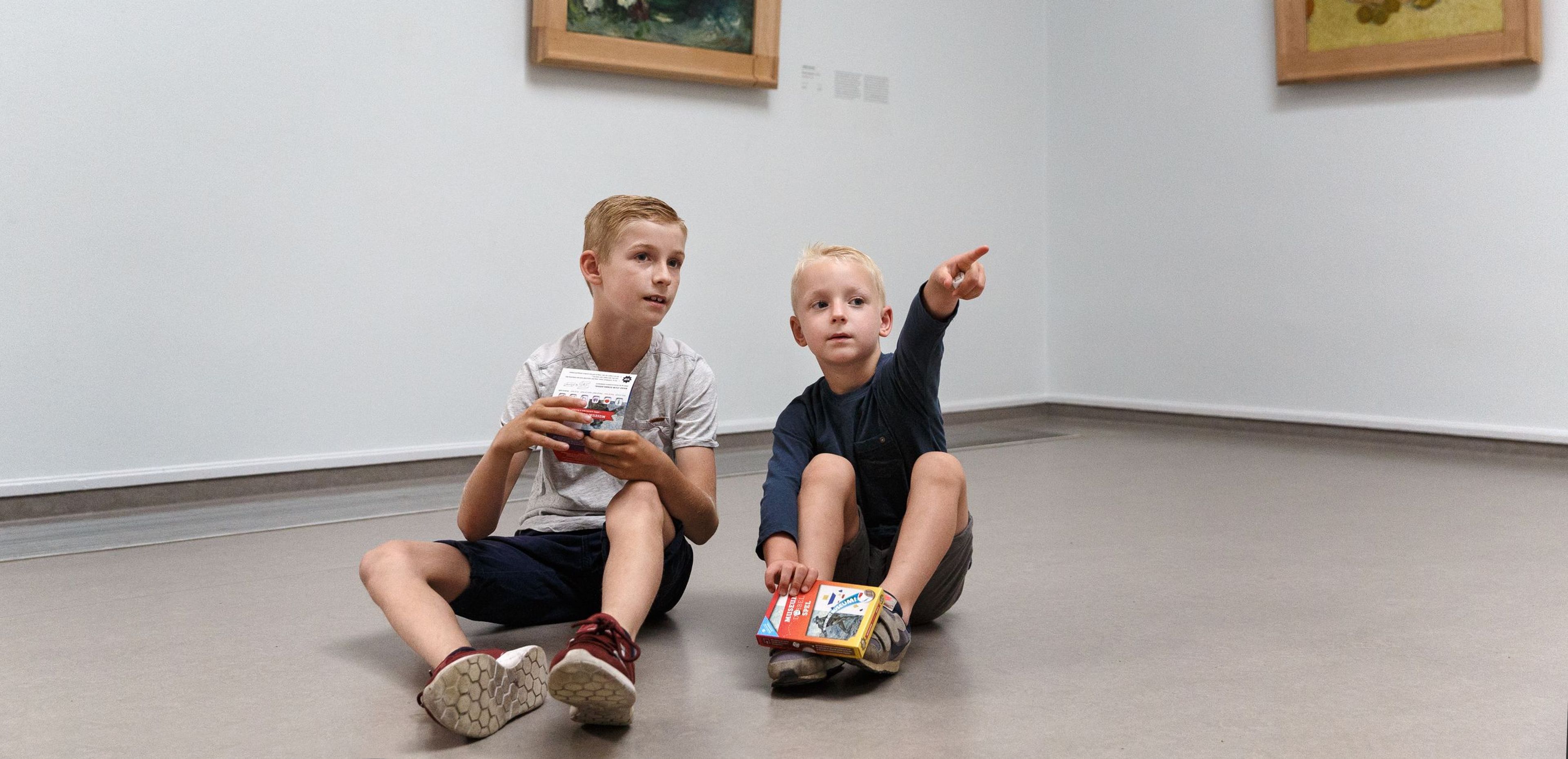 Photo of two boys sitting on the floor in a museum gallery with white walls and two framed paintings on the walls. The boys are holding a toy box; one is pointing at something outside the frame. The paintings depict flowers and a bottle of fruit.