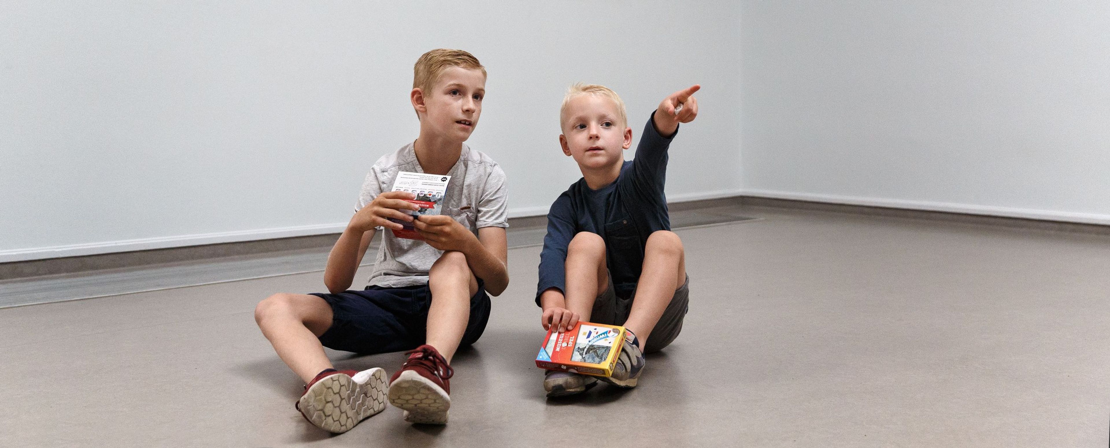Foto van twee jongens zittend op vloer in museumzaal met witte muren en twee ingelijste schilderijen aan muren. Jongens houden een spellendoosje vast, één wijst naar iets buiten beeld, schilderijen tonen bloemen en een fles met vruchten.