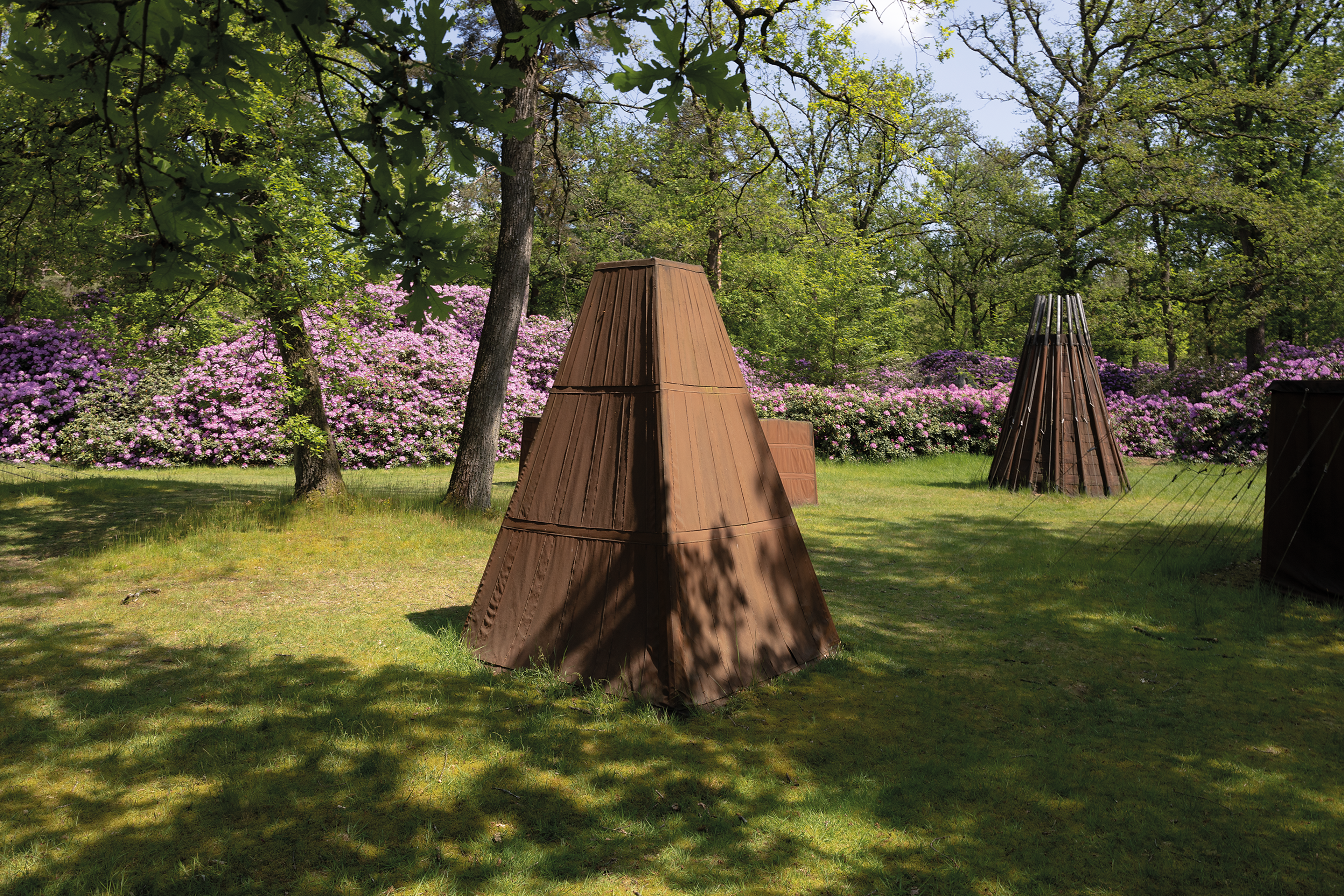 Cornelius Rogge, _Tentenproject_, 1975 Foto van meerdere bruine tenten geplaatst op grasveld omringd door bomen en bloeiende struiken.