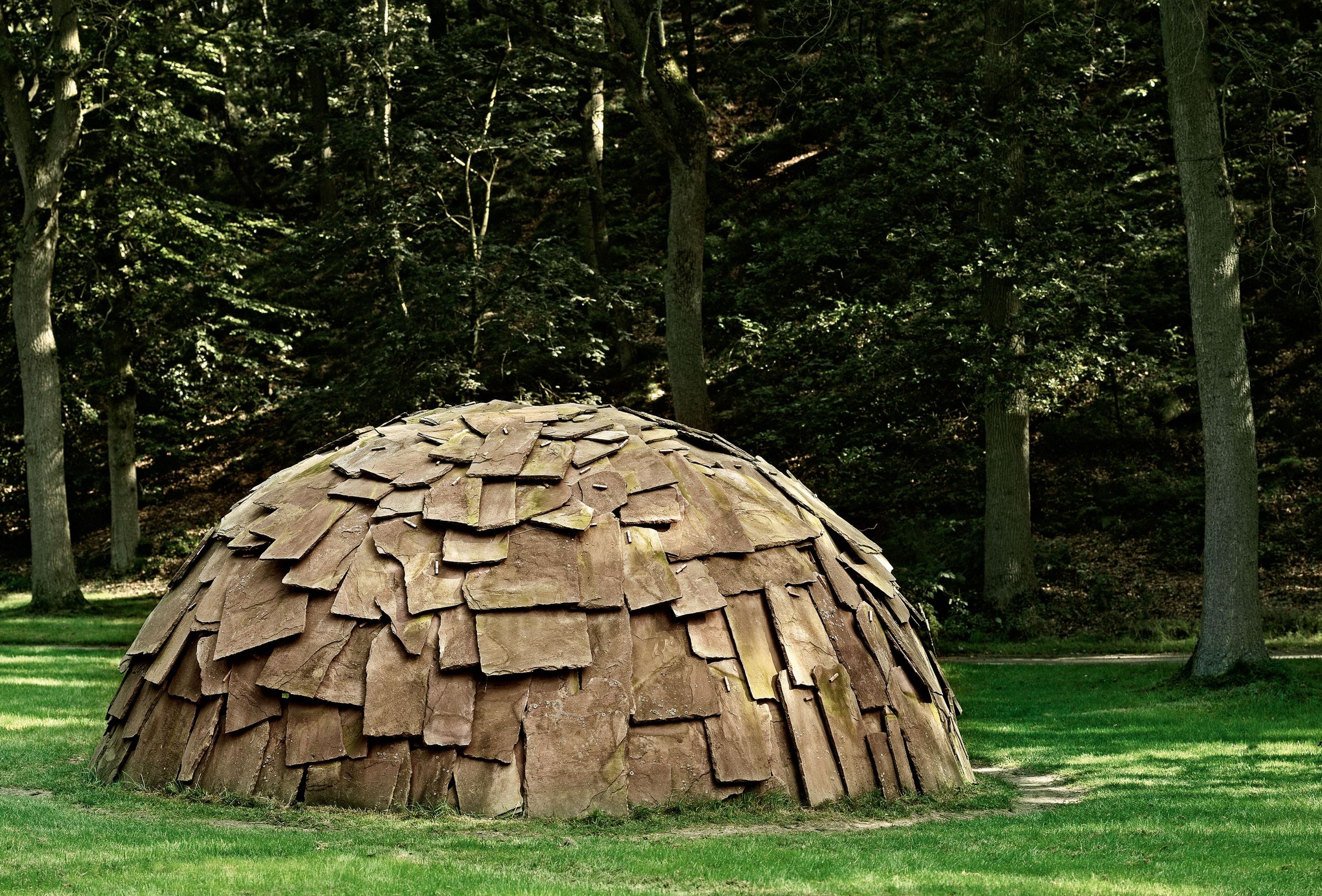 Mario Merz, (Milaan 1925- Milaan 2003), _Igloo di Pietra_, 1982, 200 x ø 450 cm, zandsteen en metaal, KM 113.373 Fotografie van een kunstwerk bestaande uit een halve bolvorm bedekt met overlappende stenen platen, geplaatst op grasveld omringd door bomen.