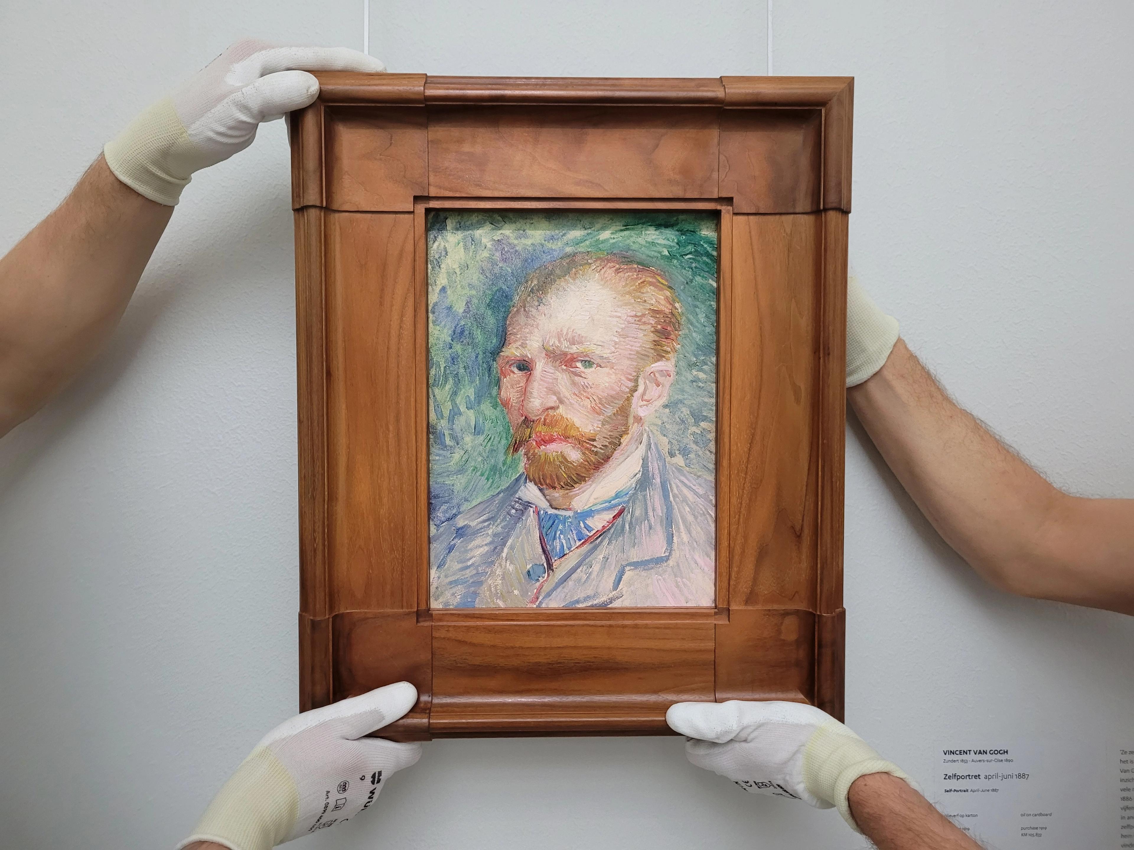 Twee handen houden Van Gogh's schilderij Zelfportret vast, en hangen het op zijn plaats