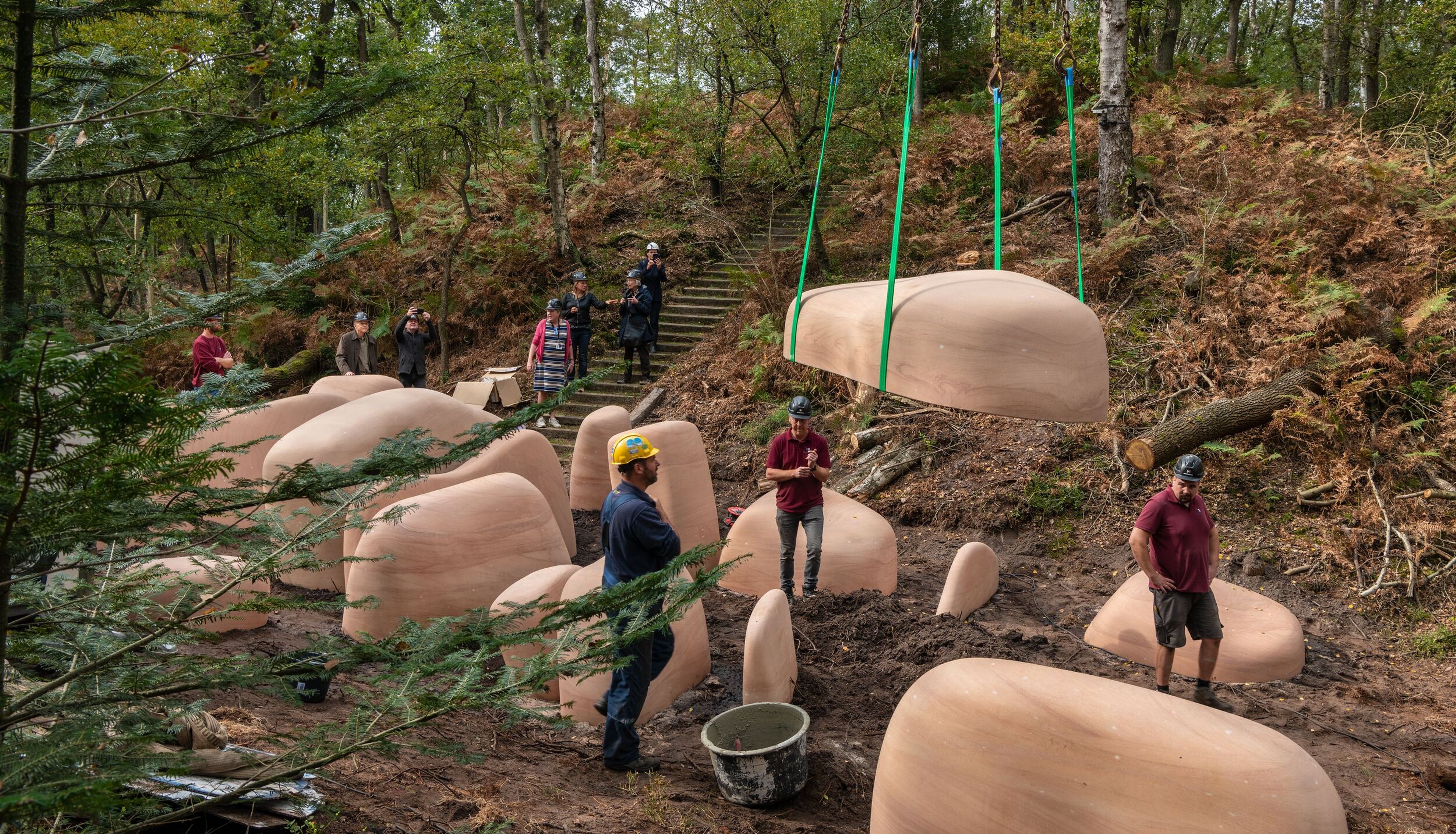 Mensen met bouwhelmen op plaatsen grote, organisch gevormde delen van een kunstwerk met hijsbanden in de bosrijke beeldentuin. Op de achtergrond kijken een paar personen toe vanaf een trap terwijl het kunstwerk wordt opgebouwd. 