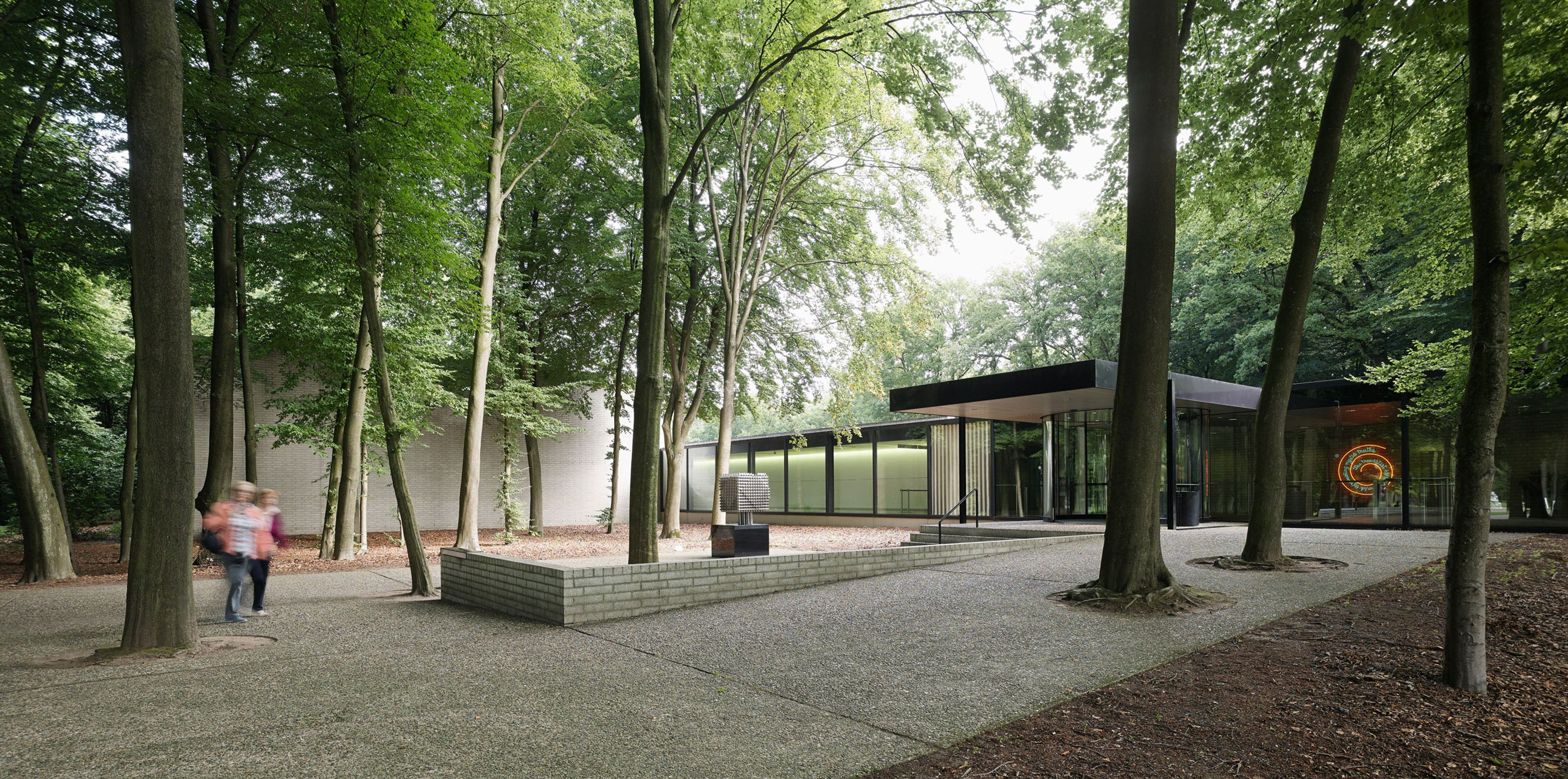 Foto van het museum omgeven door hoge bomen in een bosrijke omgeving, met een paar mensen die langs lopen. Gebouw heeft strakke lijnen en een plat da