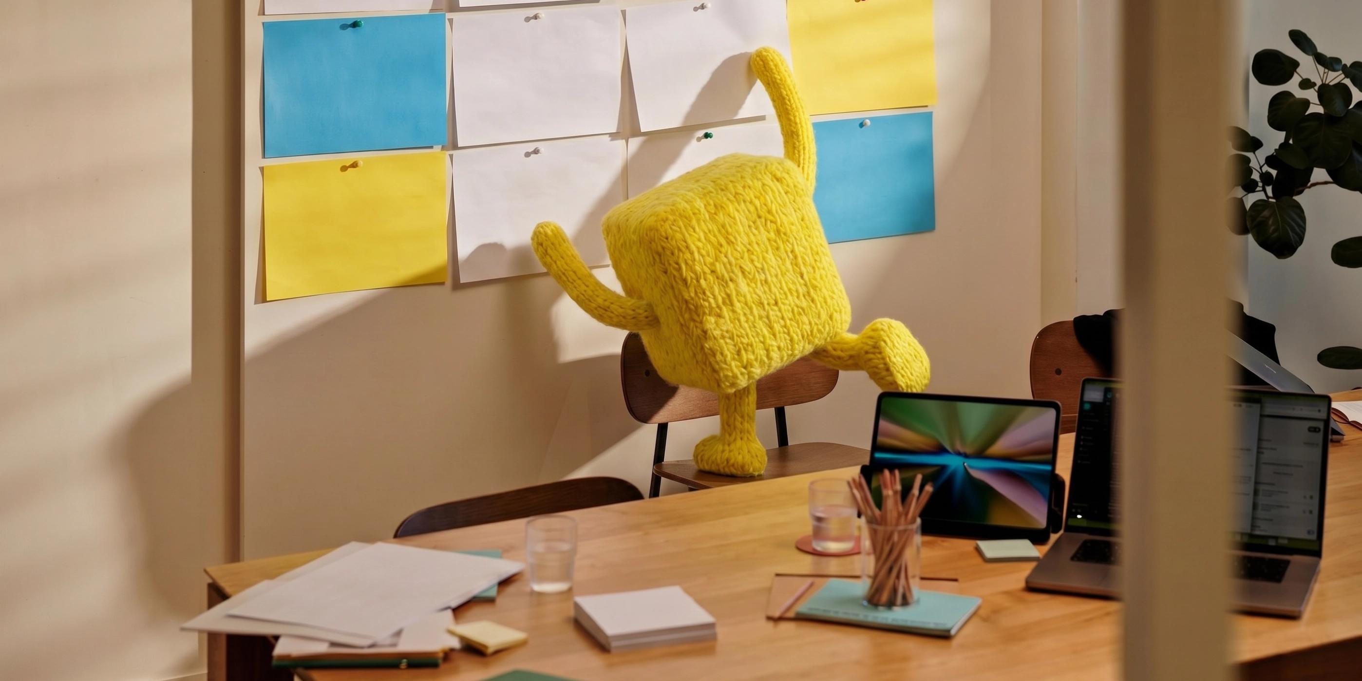 Garba's knitted yellow mascot mid-leap across a desk in the Malmö office, a wall of sticky notes behind.