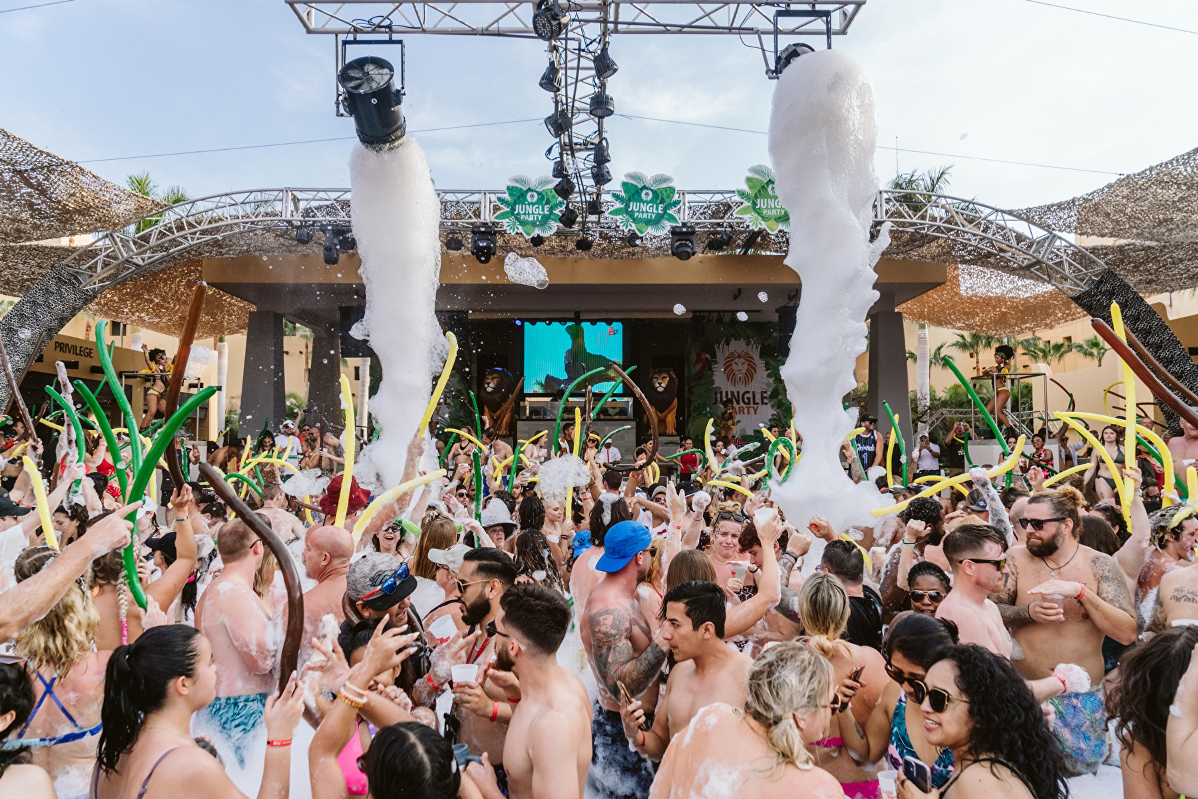 A crowd dances under foam cannons in front of the stage at a Jungle Party, around the pool