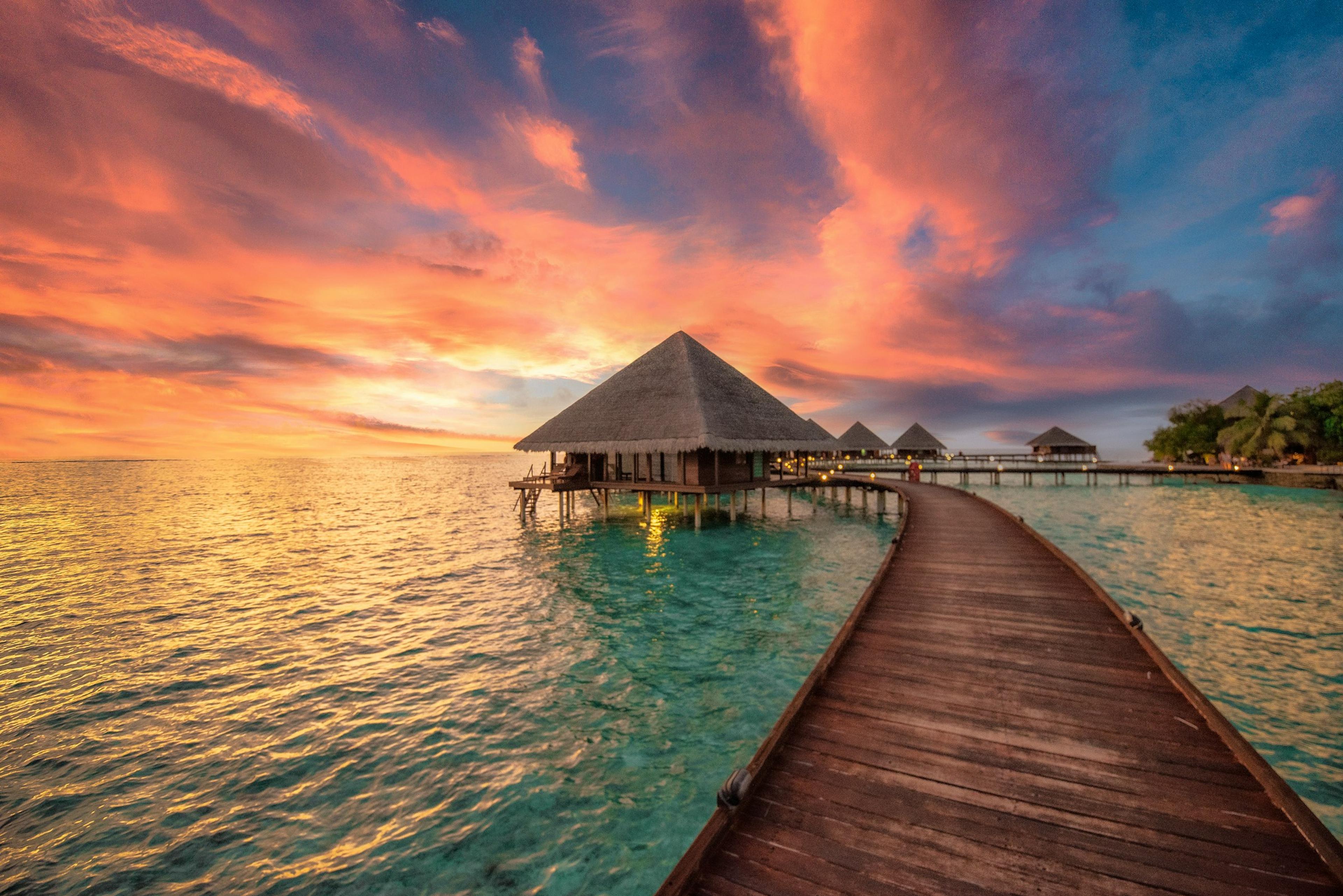 Une passerelle de bois mène à des bungalows sur pilotis, au coucher du soleil