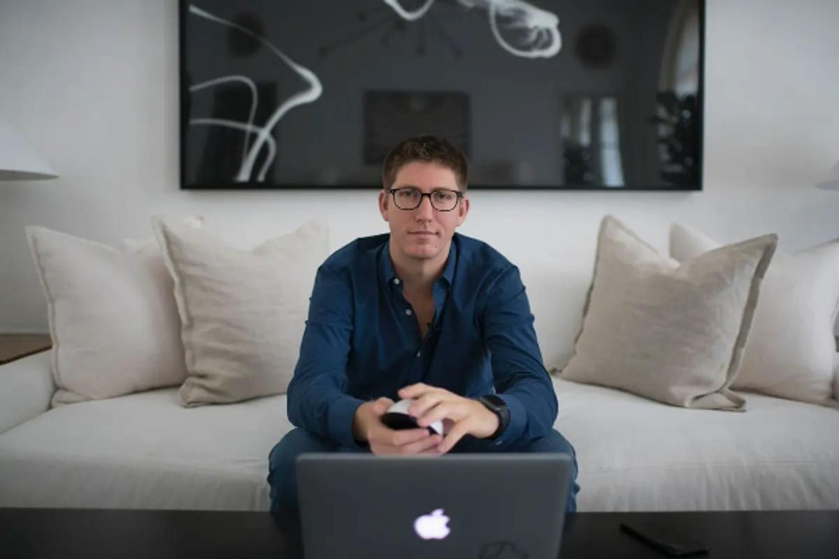Alex Capecelatro sitting on a white couch with a laptop in front of him, wearing a dark blue shirt and glasses, holding a smart remote