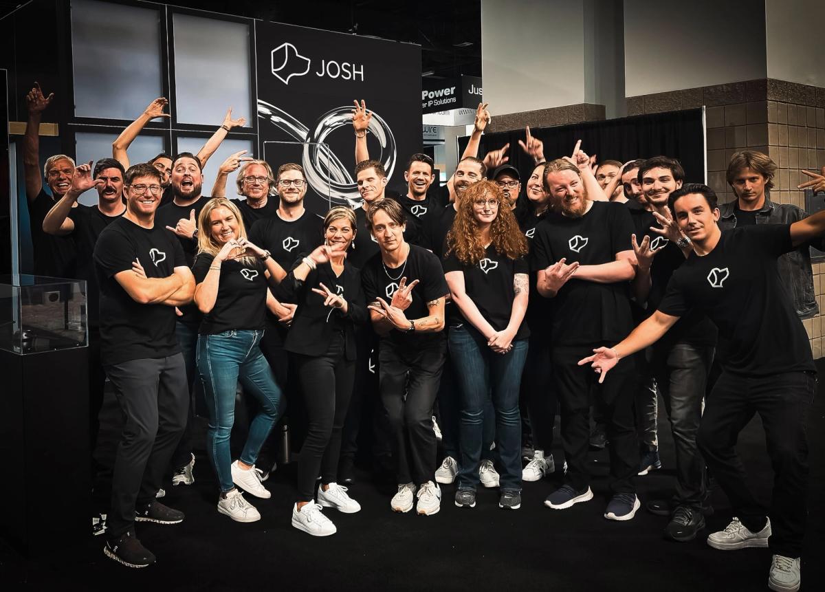 The Josh.ai team celebrating together at a trade show booth, all wearing matching black Josh.ai T-shirts and striking playful poses.