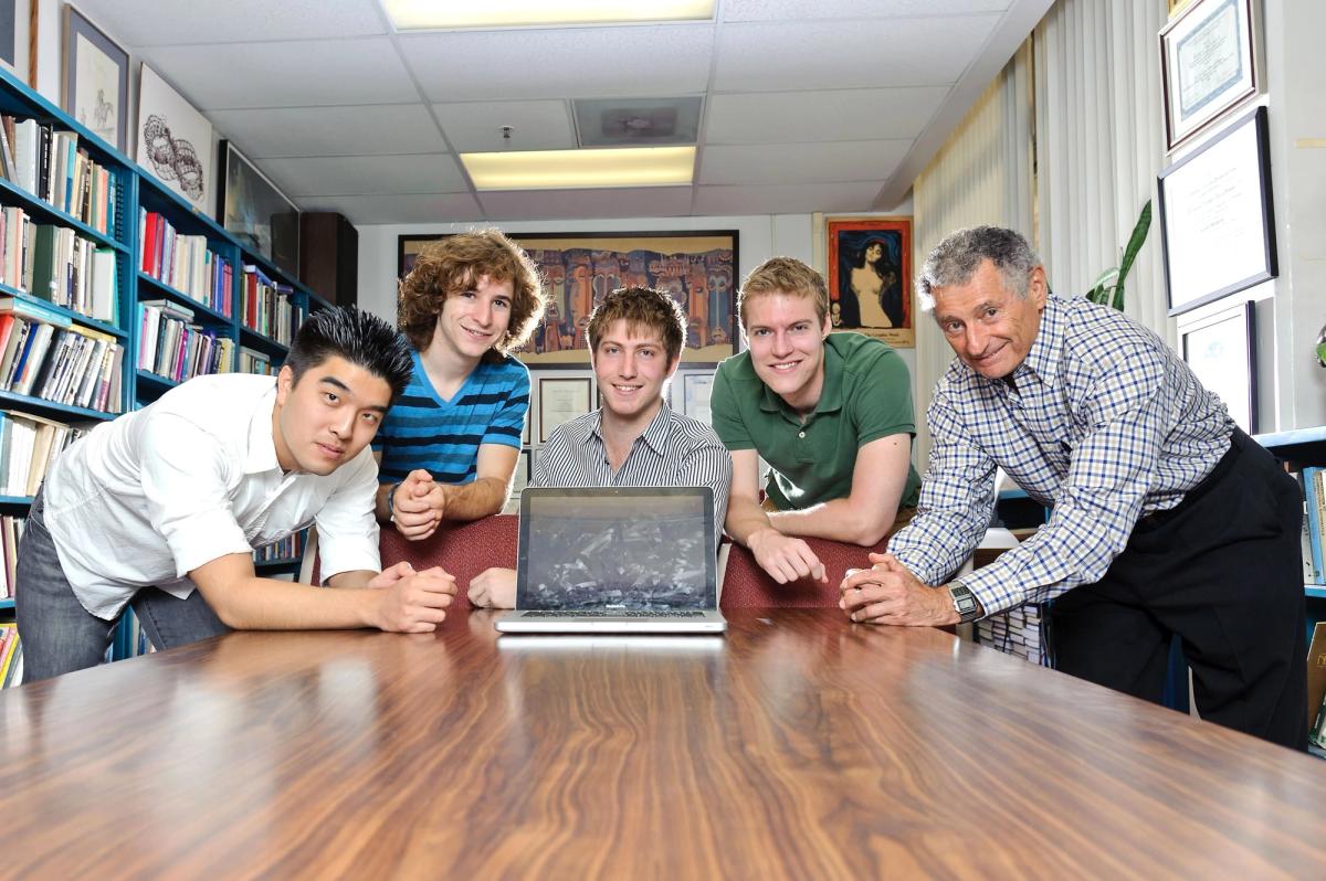 A group of young researchers posing in a university office with bookshelves and certificates, centered around a laptop on a conference table.
