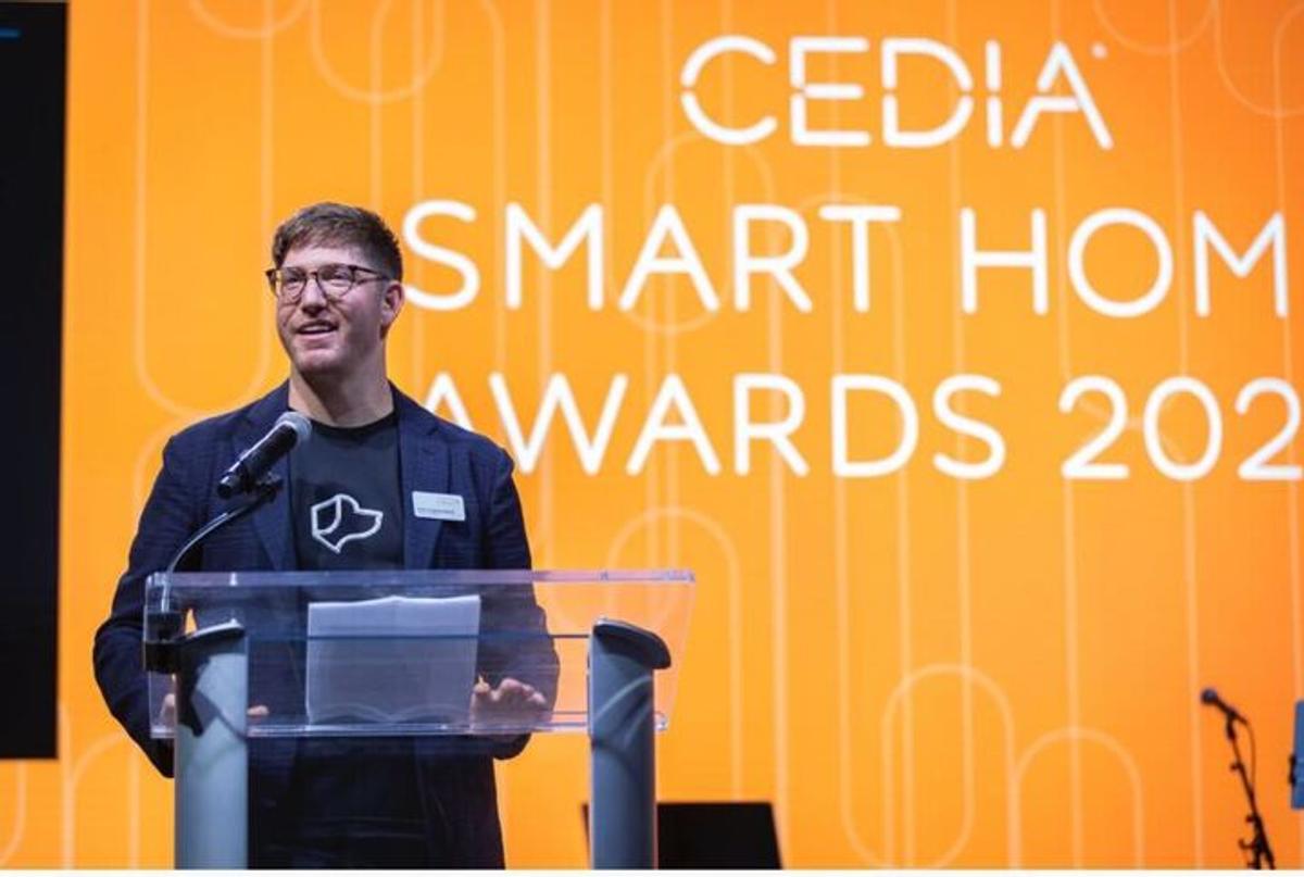 Alex Capecelatro speaking at the CEDIA Smart Home Awards 2023, standing at a podium with an orange event backdrop behind him.