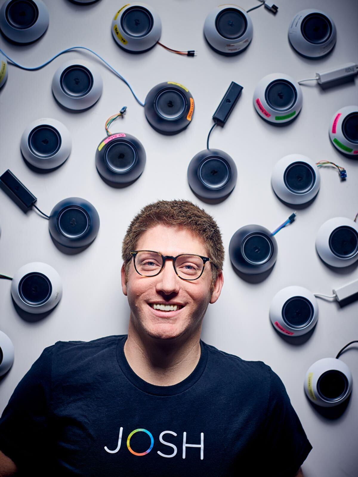 Alex Capecelatro smiling in front of a wall covered in Josh Micro devices, wearing a black Josh.ai T-shirt and glasses.