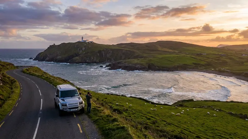 Modern commercial car on a scenic coastal road in Ireland, overlooking a bay with green hills and a lighthouse.