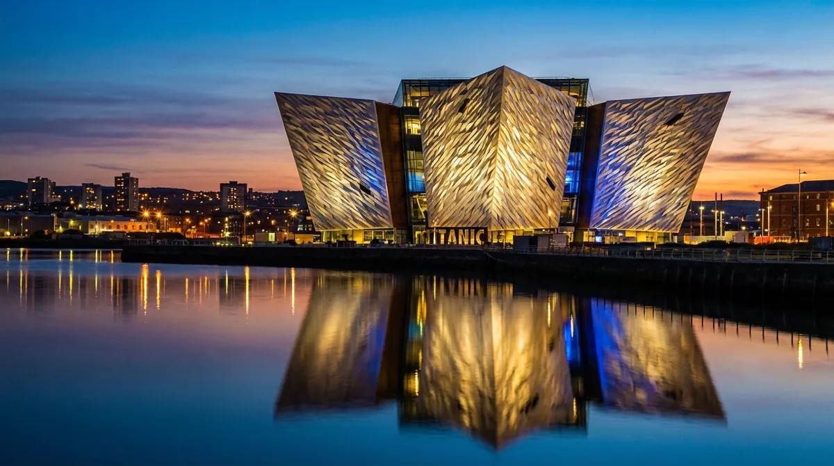 The illuminated Titanic Belfast museum building at dusk, reflecting in the water.