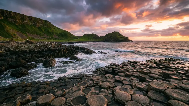 Dramatic view of the Giant's Causeway basalt columns at sunset, with waves crashing against the rocks and green cliffs in the distance.