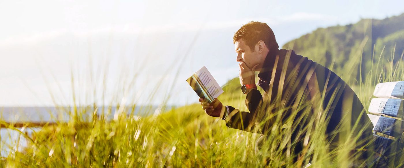 A man reads a book while sitting in tall grass near the coast