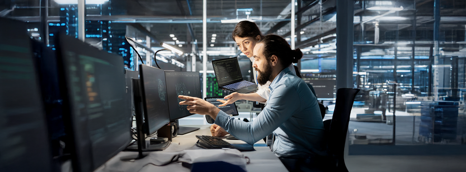 Two engineers reviewing dashboards and code across multiple monitors in a data center