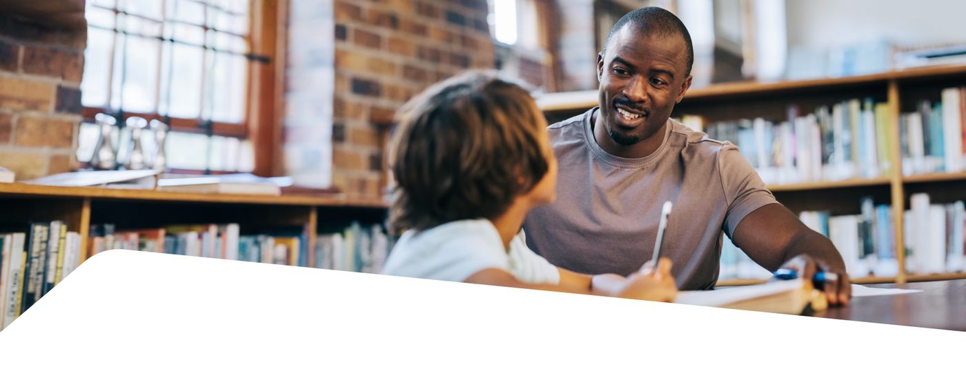 A tutor helps a young student write at a table in a library