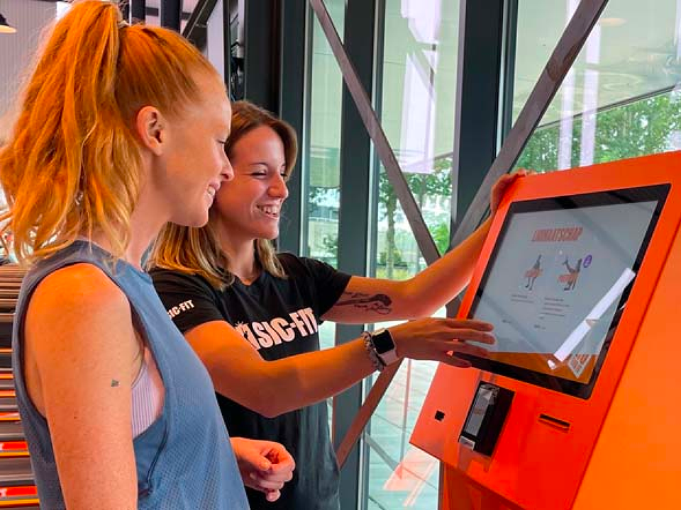 Two members using a Basic-Fit self-service kiosk to select a workout