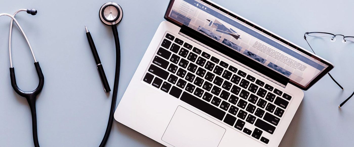 A stethoscope, glasses and pen laid out beside an open laptop showing a website