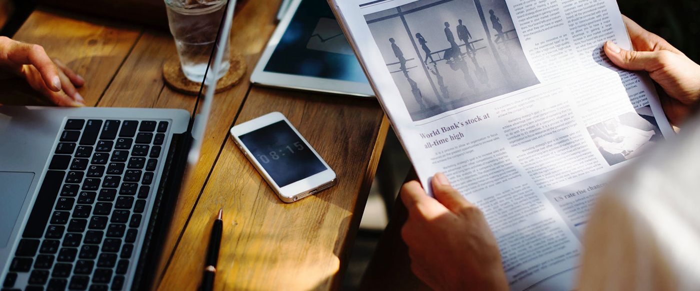 Hands hold a newspaper reading 'World Bank's stock at all-time high' beside a laptop and phone