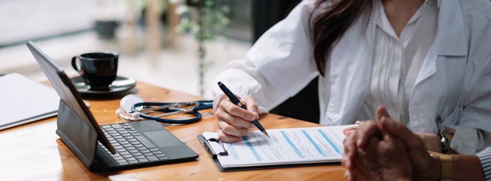 Doctor writing on a clipboard next to a laptop and stethoscope on a desk