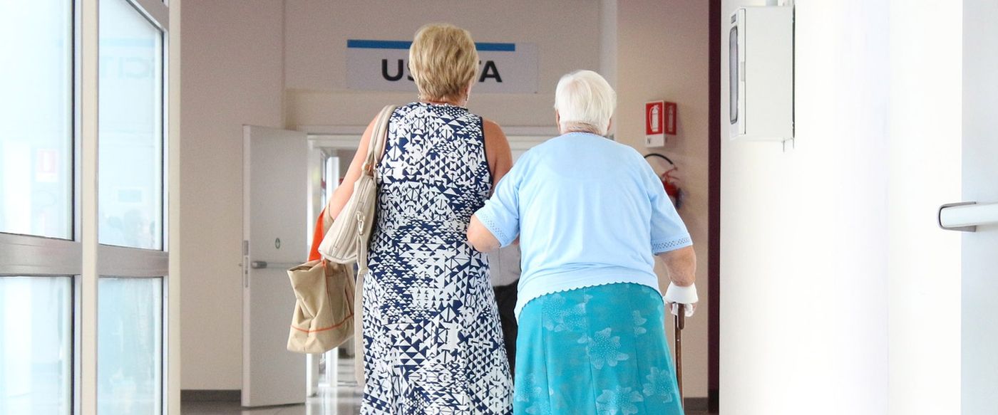 An older patient being helped down a hospital corridor by a companion
