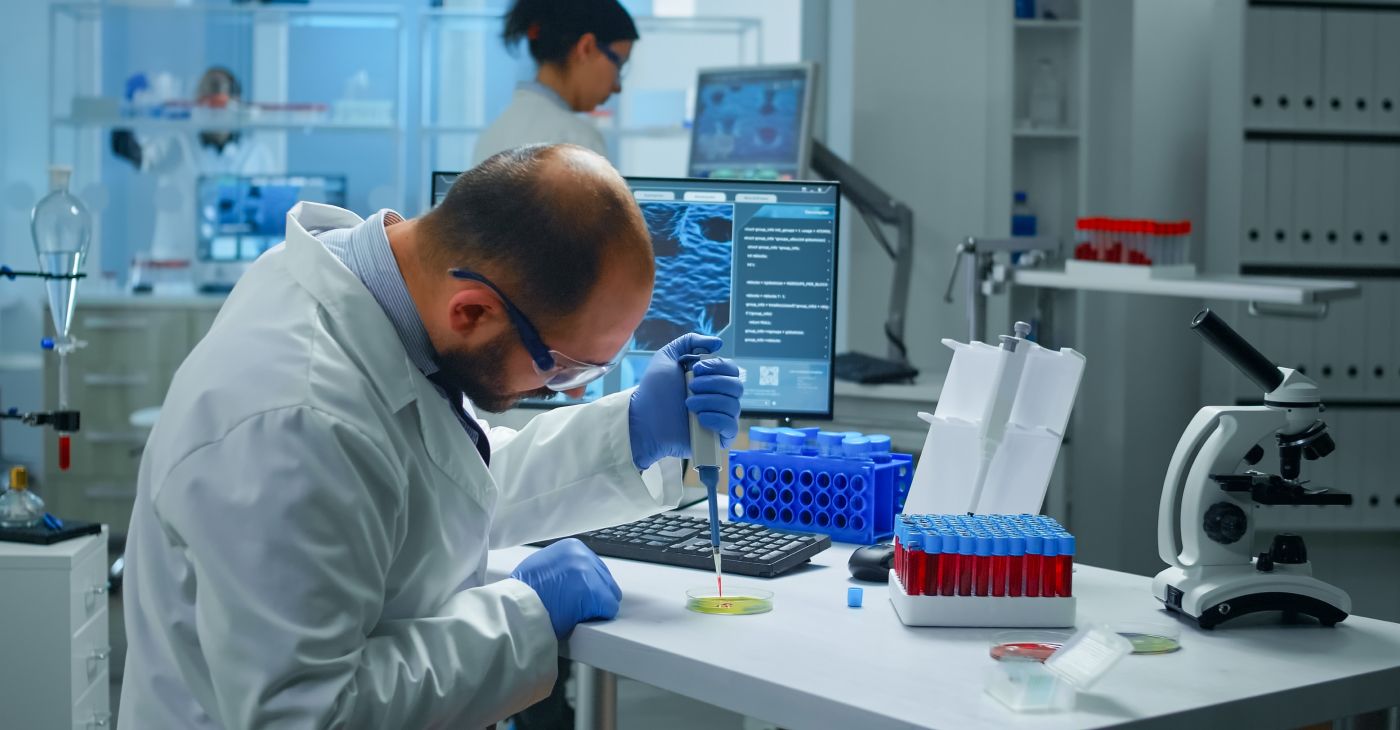 A lab scientist using a pipette at a workstation with blood sample tubes and a microscope