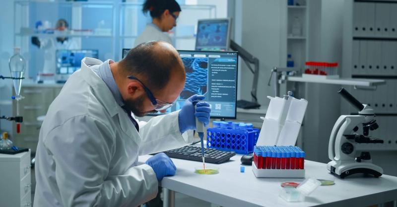 A lab scientist using a pipette at a workstation with blood sample tubes and a microscope