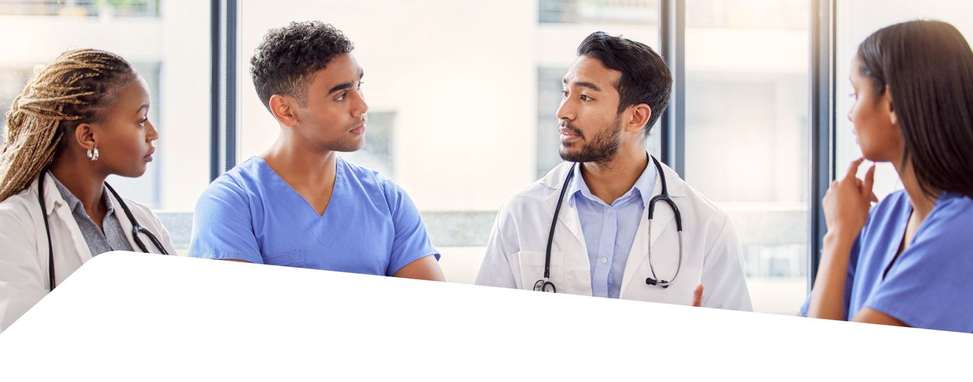 Four healthcare professionals in scrubs and white coats converse in a meeting