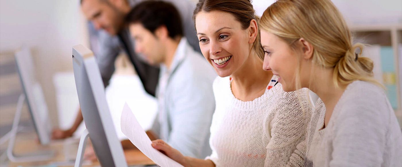 Two colleagues smile while looking together at a computer screen in an office