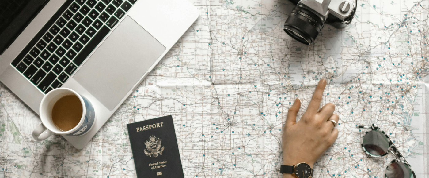 A flat-lay of a laptop, passport, coffee and map with a hand pointing to a route