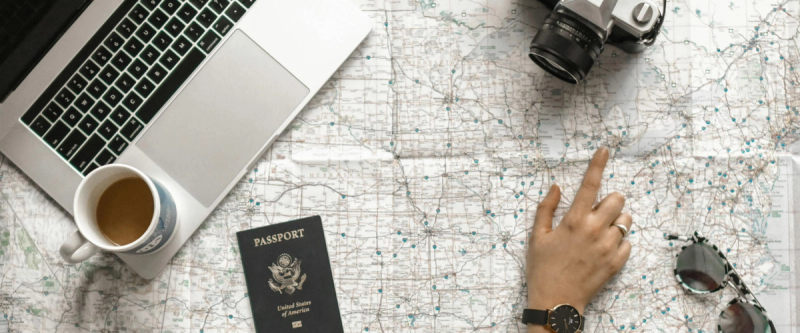 A flat-lay of a laptop, passport, coffee and map with a hand pointing to a route