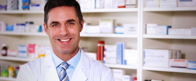 A smiling pharmacist stands in front of shelves stocked with medication