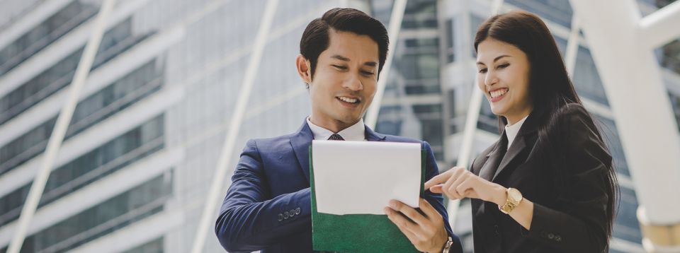 Two businesspeople review notes on a tablet outside a modern office building