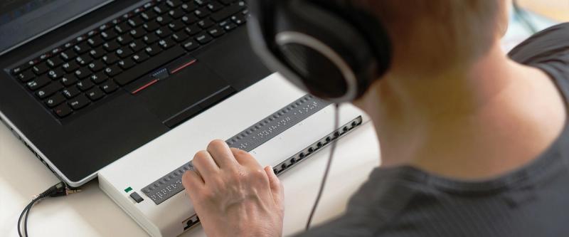 A person uses a refreshable braille display and headphones alongside a laptop