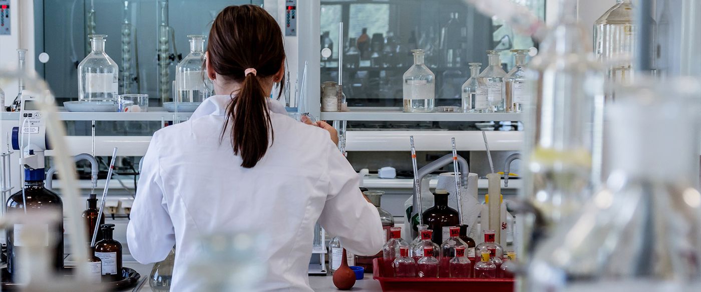 A lab scientist in a white coat works among rows of laboratory bottles