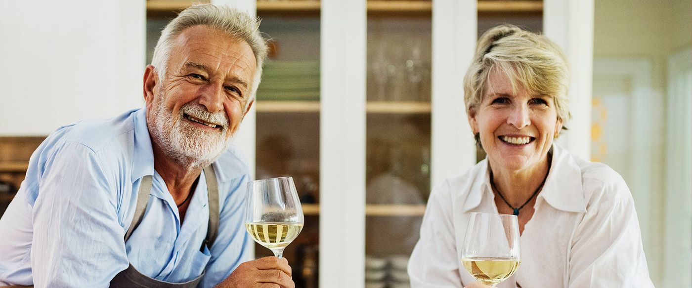 An older couple smile together while holding glasses of white wine in a kitchen