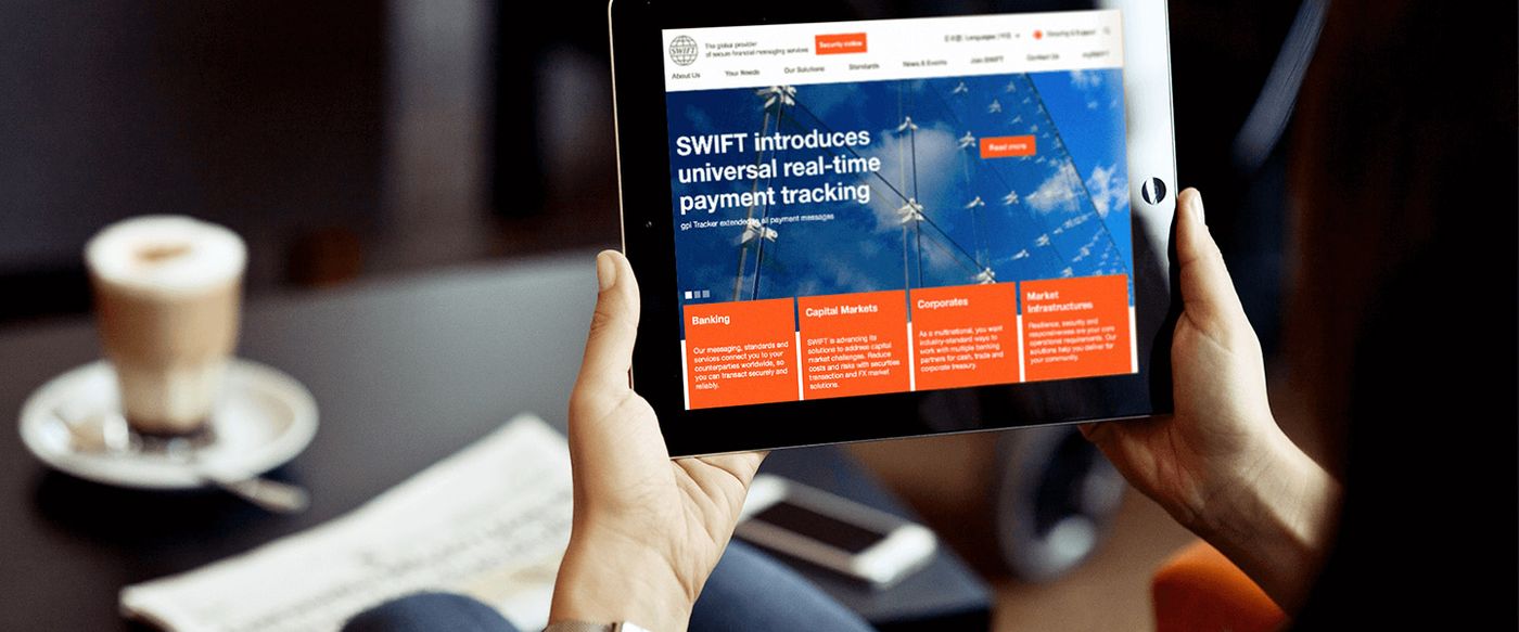 Hands holding a tablet showing the SWIFT website announcing universal real-time payment tracking