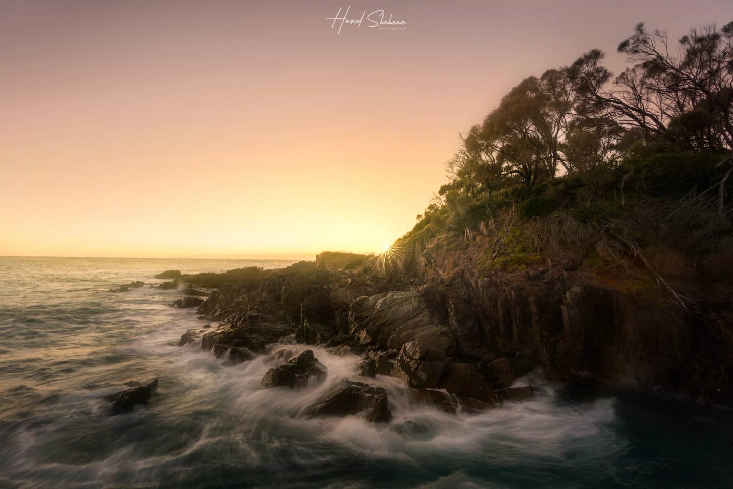 Waves flowing around a rocky, tree-covered headland at sunset.