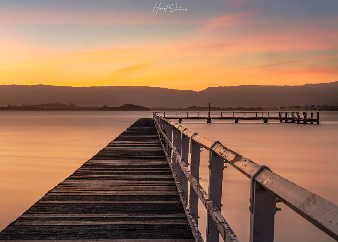 A long timber jetty extending across still water towards distant hills at sunset.