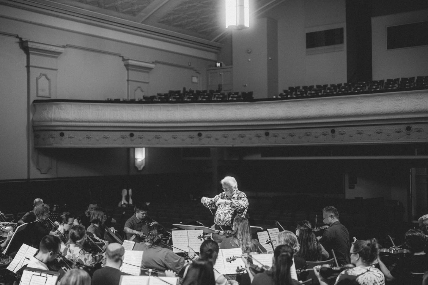 A conductor leading seated orchestral musicians beneath the concert hall balcony, in black and white. Australian Doctors Orchestra 2026, Adelaide.