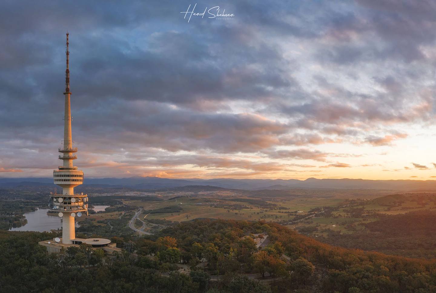 Telstra Tower viewed from the air above forested hills beneath a glowing evening sky.