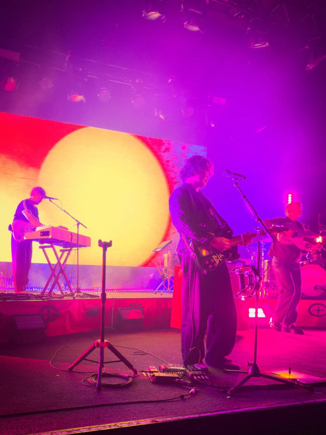 A band performing under purple and magenta stage lights, with an electric guitarist in front of a glowing yellow backdrop.