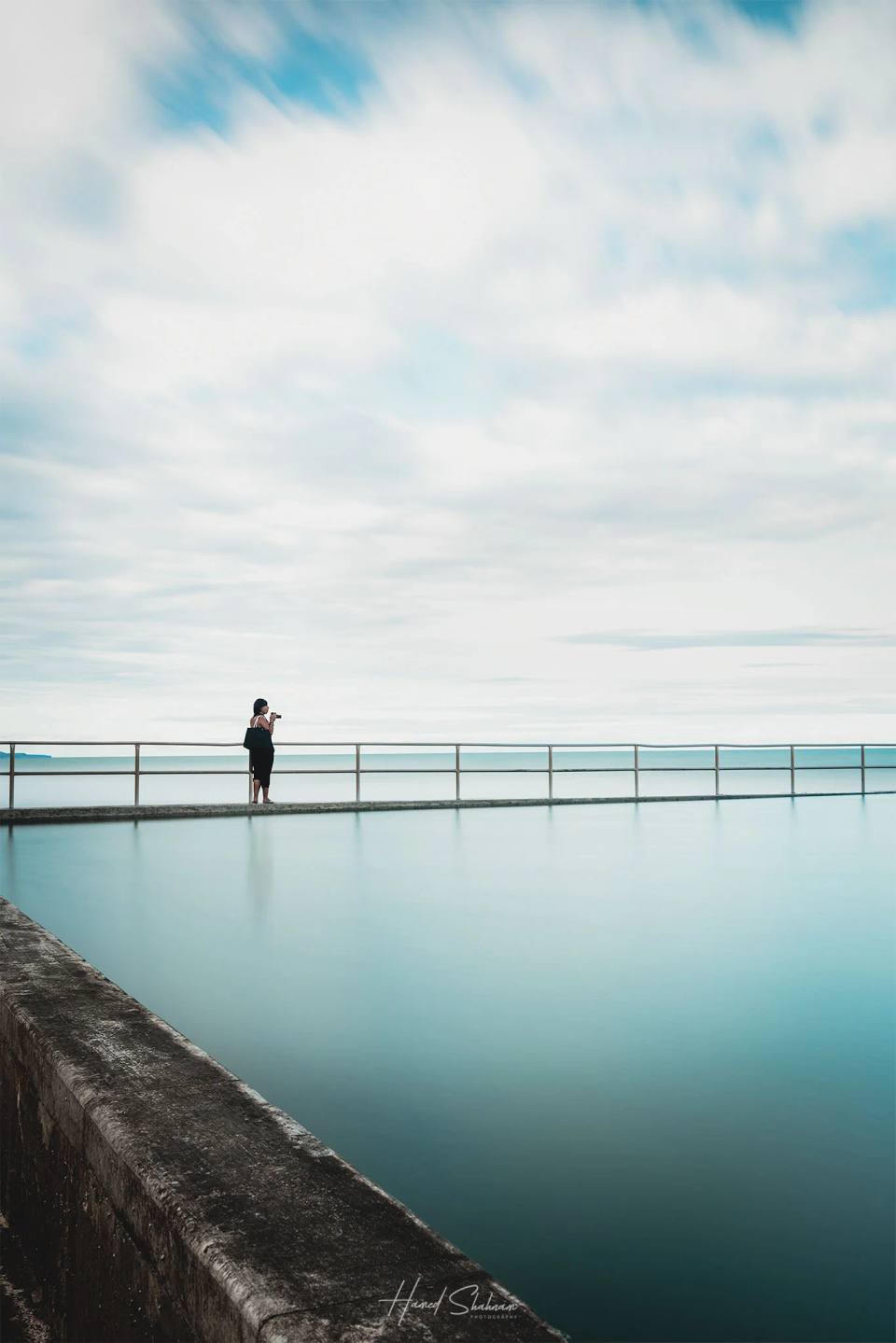 A lone person beside a calm ocean rock pool beneath a cloud-filled sky.