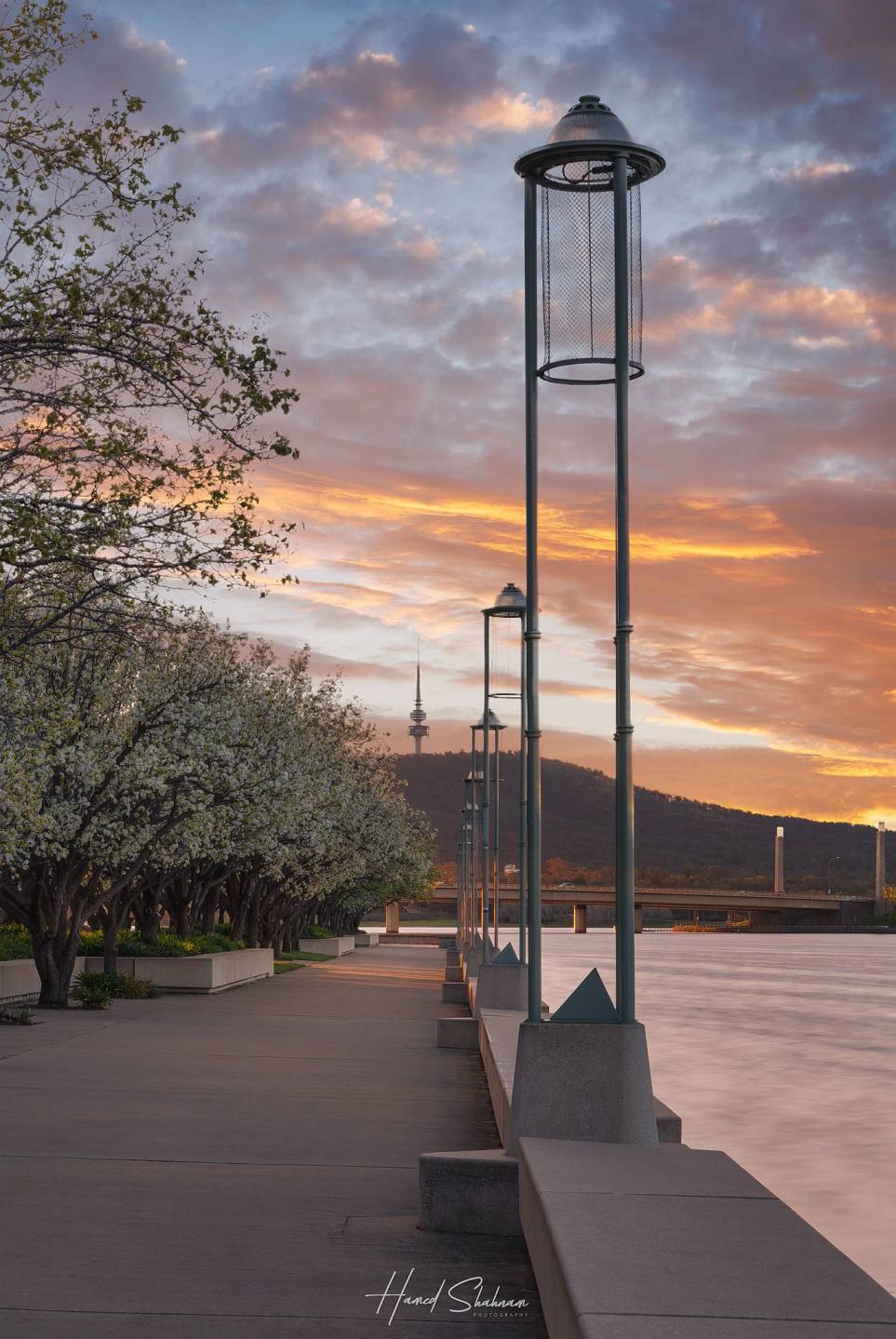 A slender metal structure beside a lakeside walkway beneath a colourful sunset.