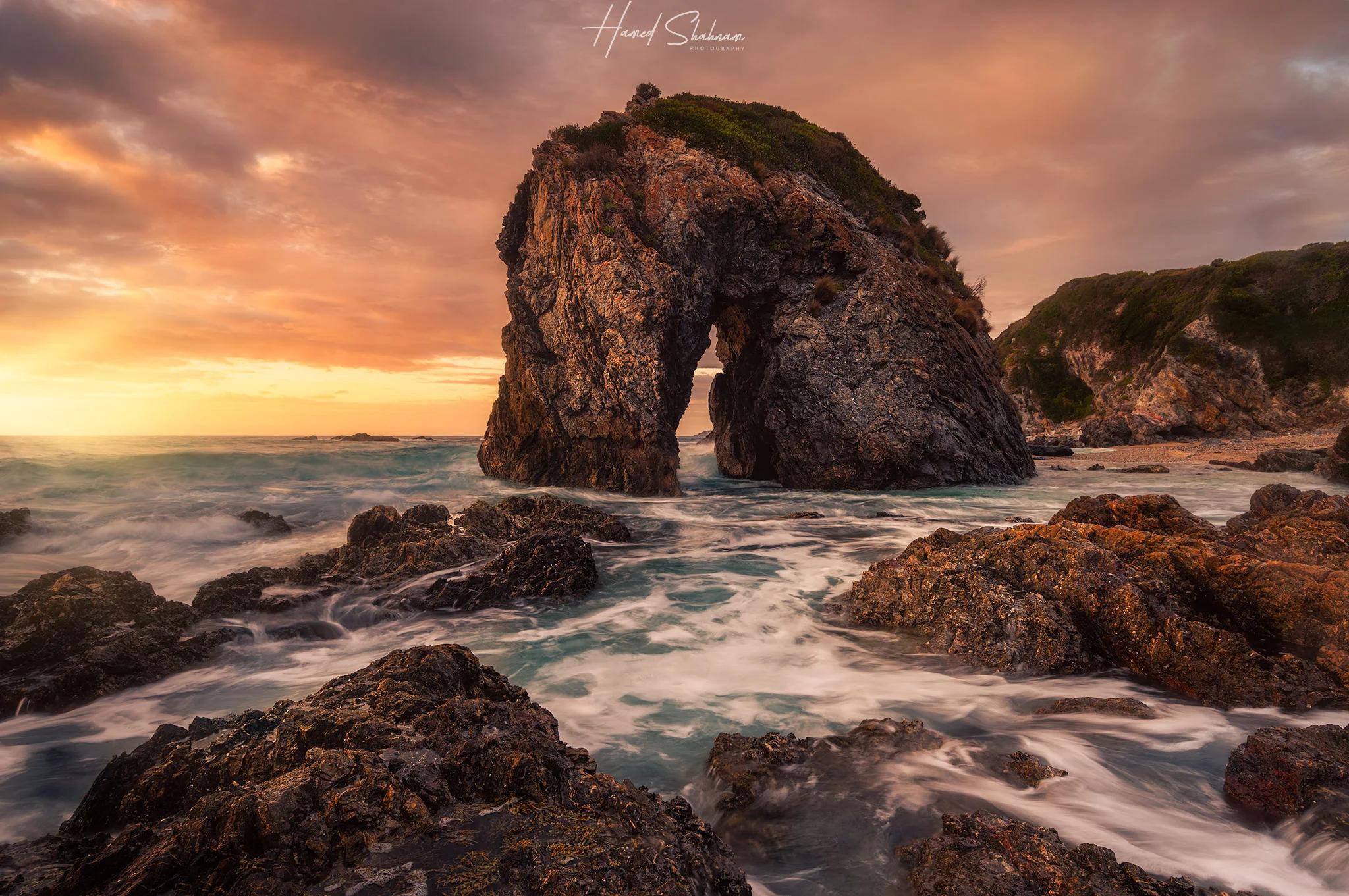 A coastal rock arch and waves beneath a glowing sunset sky.