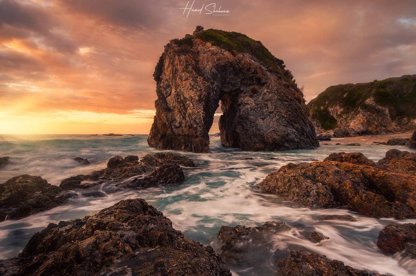 A coastal rock arch and waves beneath a glowing sunset sky.