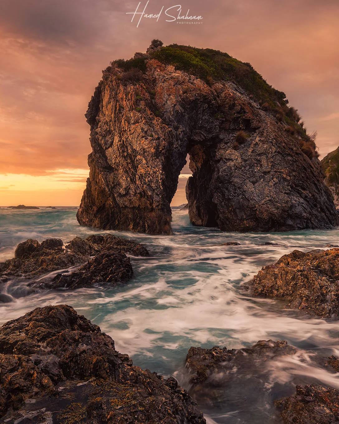 A coastal rock arch and waves beneath a glowing sunset sky.